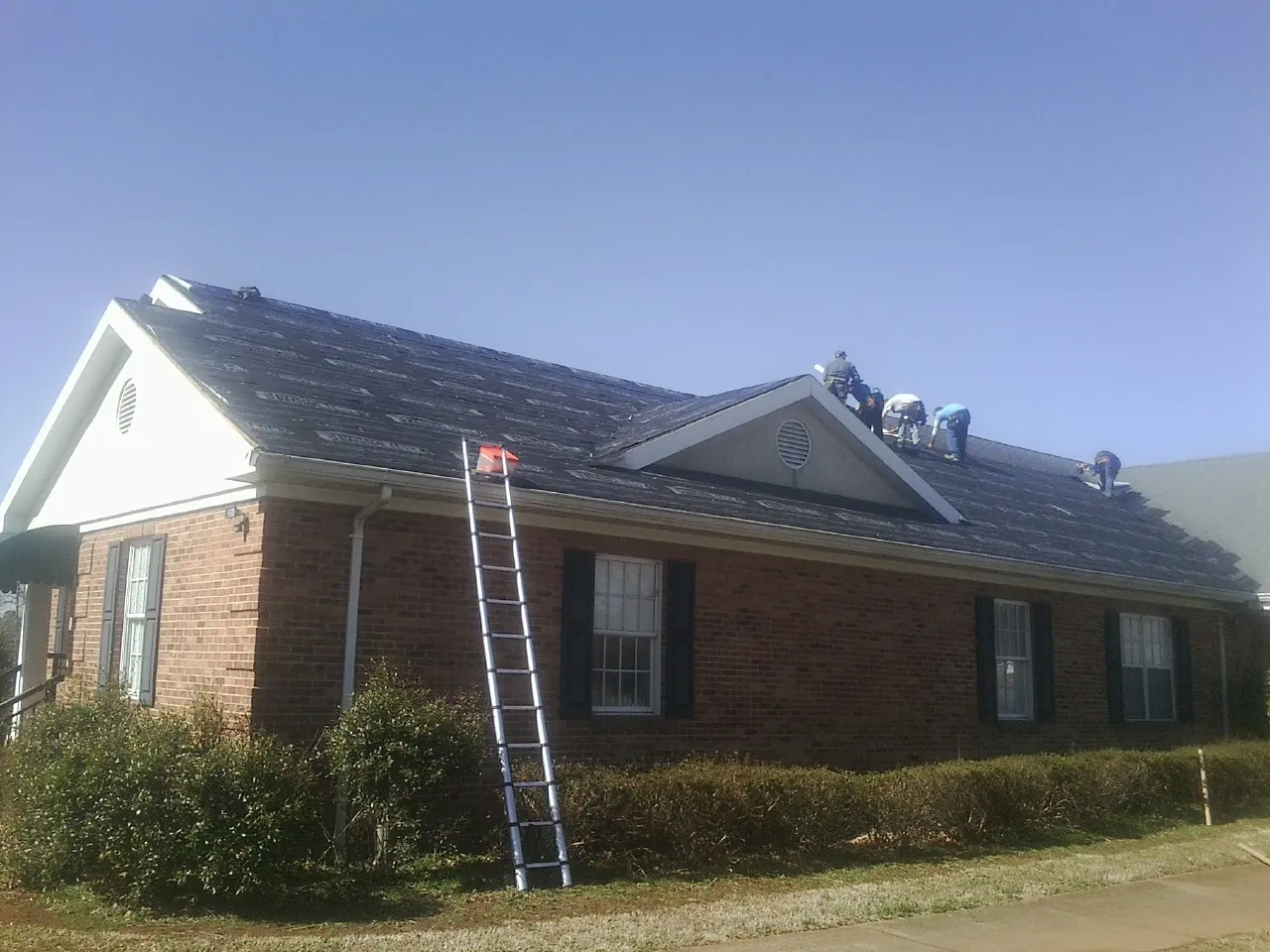 Crew tearing shingles off a brick ranch roof with a ladder set — Metal Roof Repair work by our crew in Sylvia