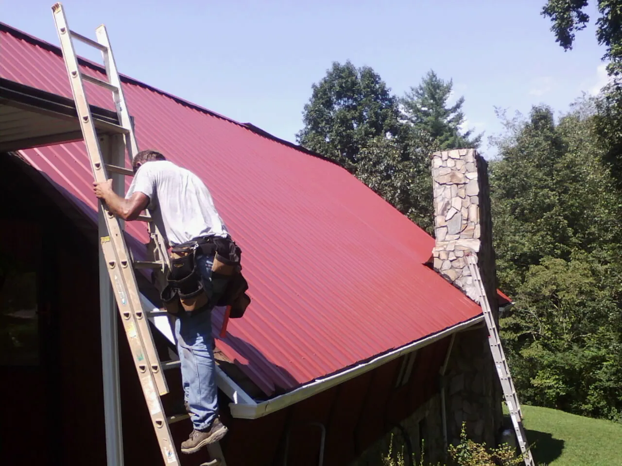 Roofer on a ladder beside a red metal roof and stone chimney — Soffit & Fascia Repair by Roofing Brothers in Sylvia